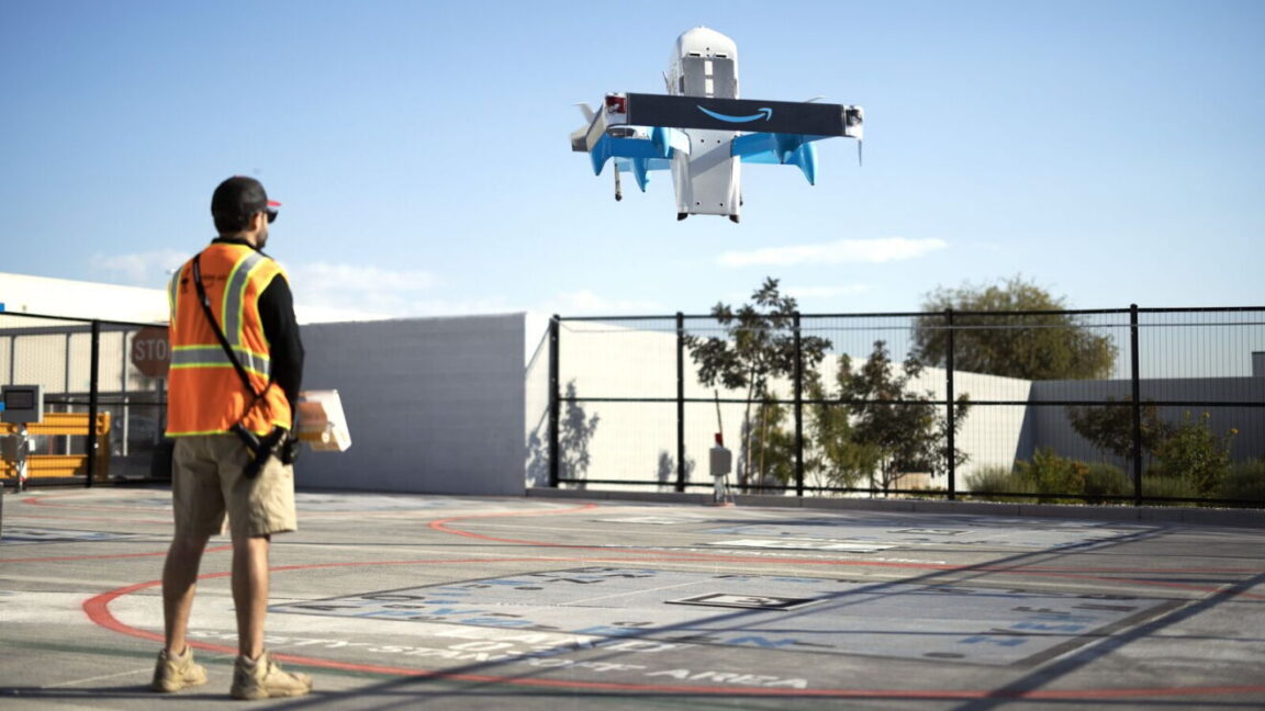 Дрон Amazon Prime Air завис над асфальтованою ділянкою перед людиною в помаранчевому сигнальному жилеті.